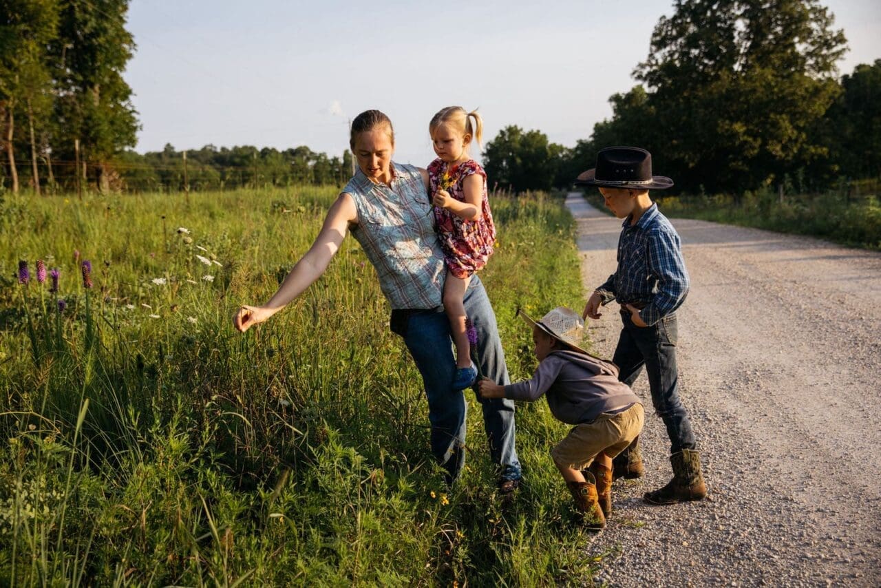 Toxic grass:A blond woman holding a baby as she leans over to pick a flower from a field of tall grass. Two children beside here.