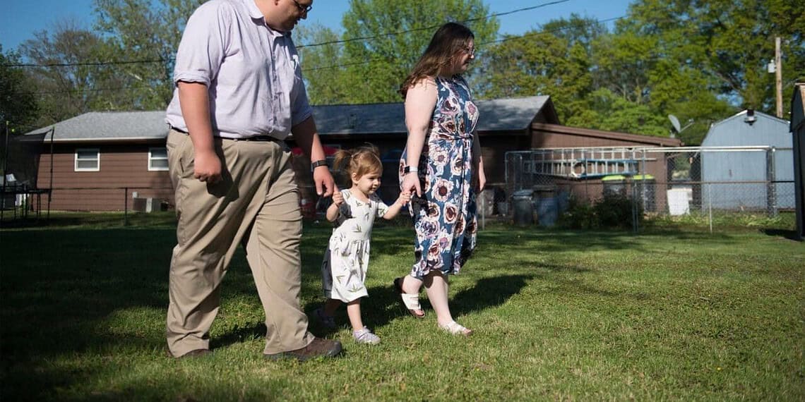 Parents holding hands with their young child between them walks through the front yard of their home