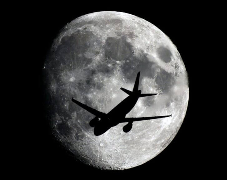 The dark silhouette of an airplane in front of a large, bright moon.