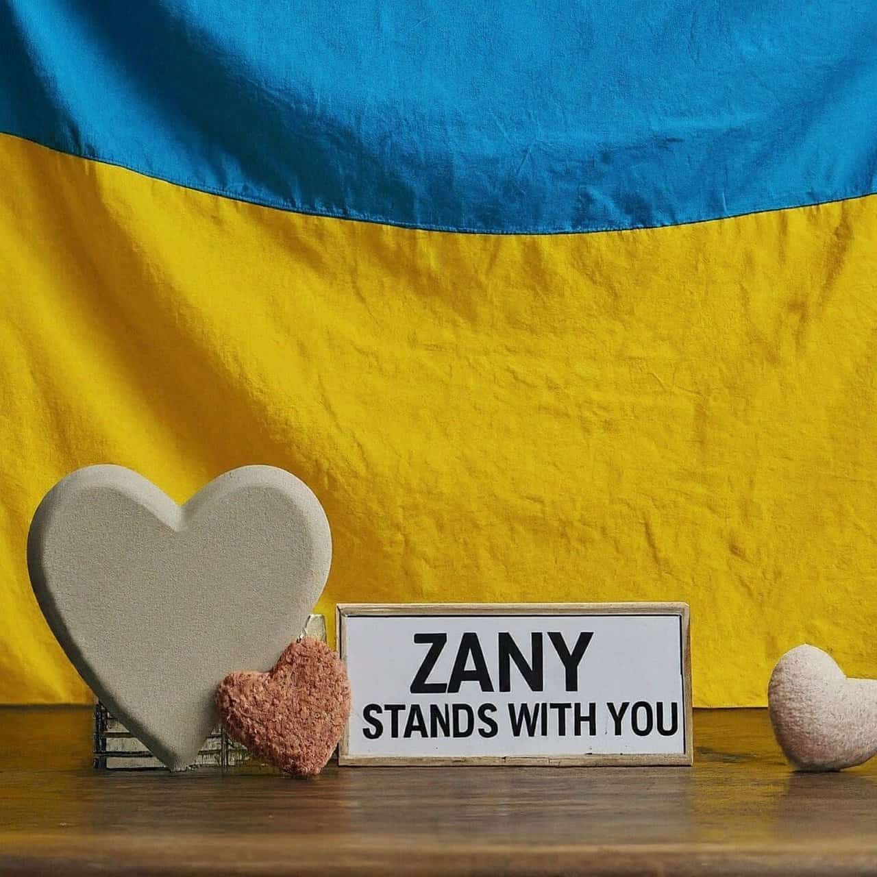 The ukrainian flag hangs in the background with a decorative heart on a wooden table in front of it along with a plaque that says “zany stands with you”