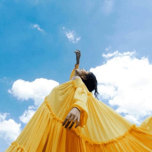 Model wearing a yellow dress points up at a bright blue sky with white fluffy clouds