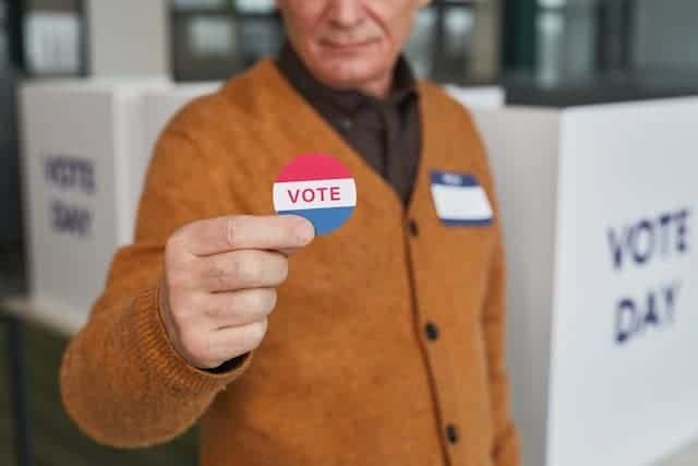 Vote against their own best interests. A man in a brown sweater holds up a round sticker that says vote