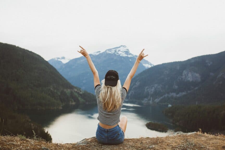 Eco-friendly lifestyle woman sitting on top of a big hill or mountain looking out over a lake making 2 peace signs with her arms raised