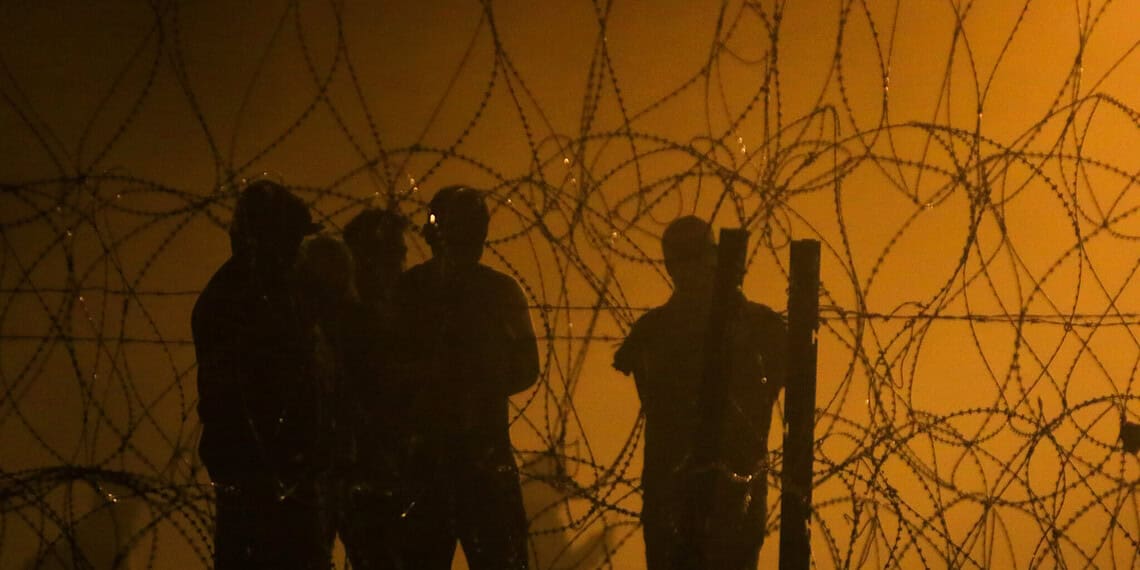 Migrants looking through a barbed wire fence as the sun rises in an orange sky