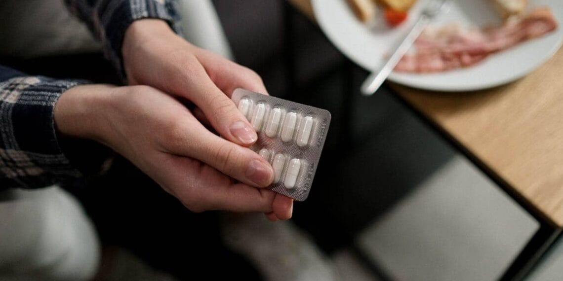 Close-up of a person holding a blister pack of pills with breakfast in the background, indicating medication and daily routine.