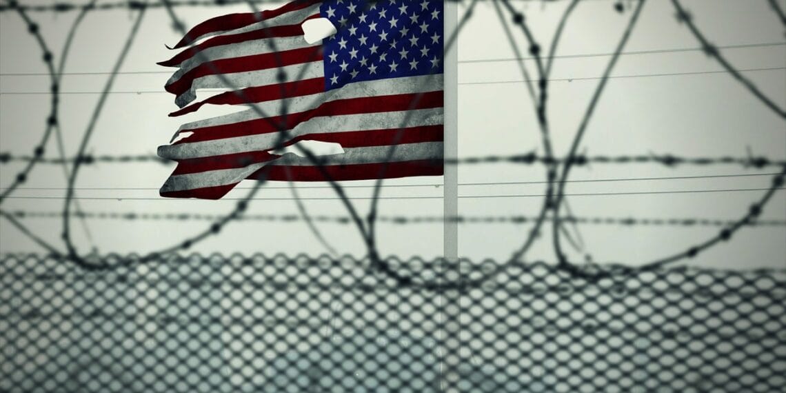 Torn american flag viewed through a fence with razor wire